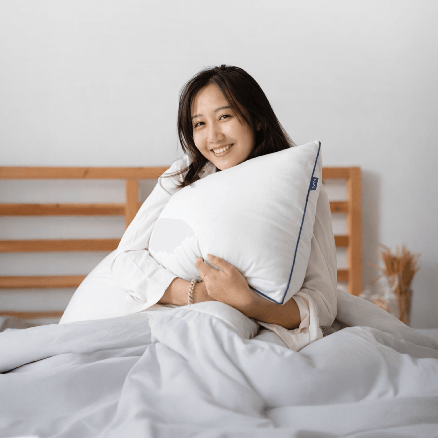 Woman holding a white Sonno Cloud pillow with a Sonno logo in a bedroom setting