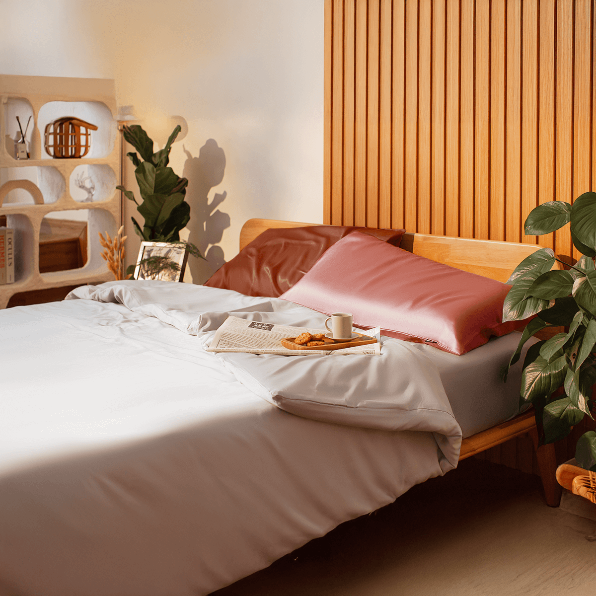 Cozy bedroom featuring a wooden bed frame with soft, neutral bedding and two decorative pillows. A small breakfast tray with a cup of coffee and a magazine rests on the bed, complemented by indoor plants and a stylish wooden accent wall. Ideal for promoting home decor and relaxation.