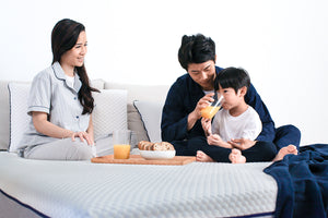 A young boy’s father helping him drink a glass of orange juice, next to his mother and a wooden tray of food, all on top of a Sonno Original mattress