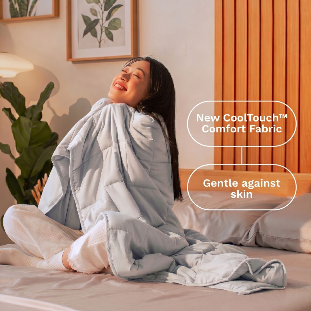 Woman sitting on a bed wrapped in a gray weighted blanket with text about 'New CoolTouch Comfort Fabric' and 'Gentle against skin'.
