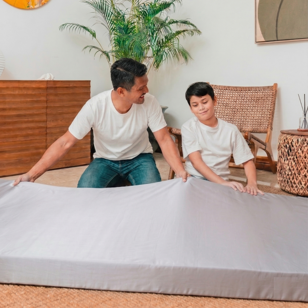 Two people putting a fitted sheet on a foldable mattress together in a room with plants and furniture.