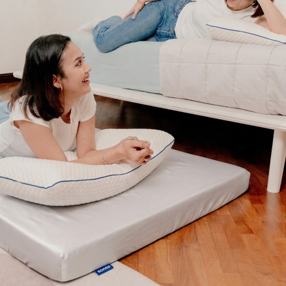 Woman lying on a foldable mattress with Tencel fitted sheet a pillow, another person sitting on a bed in the background.