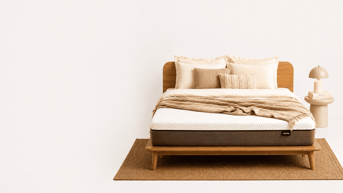 A modern Sonno Signature Bed Frame, styled with beige and white bedding, a textured throw blanket, and a Sonno Original mattress. The bed is positioned on a woven jute rug against a clean white wall, with a small side table and lamp on the right.