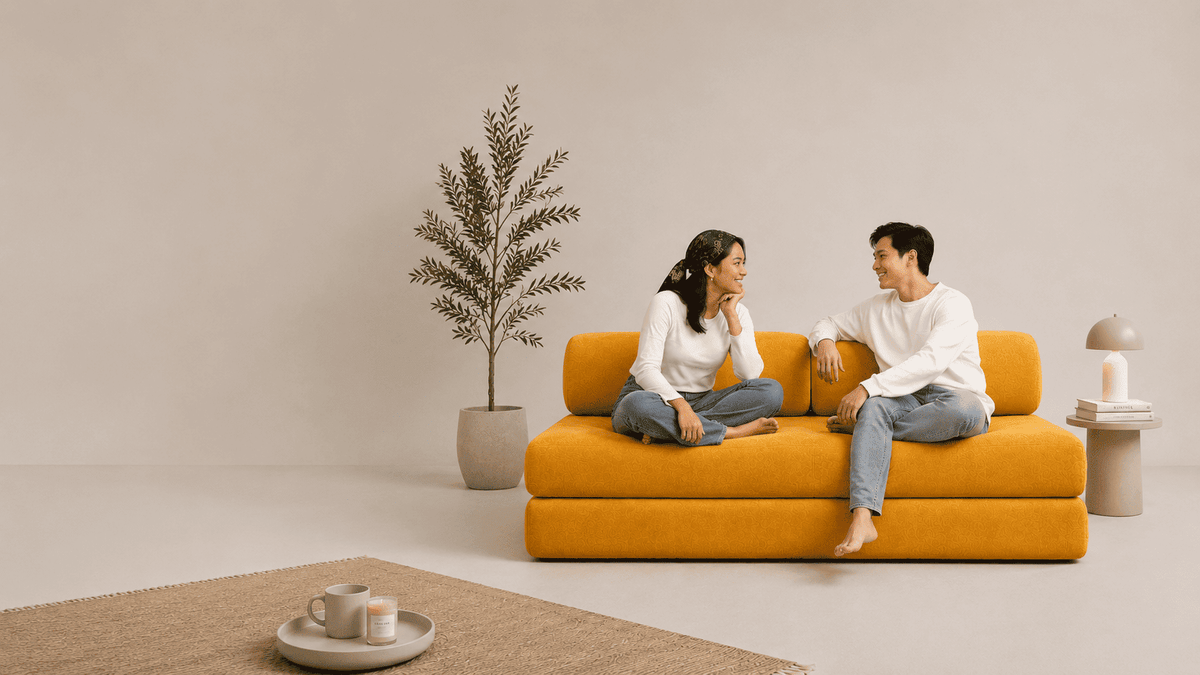 A man and a woman sit on a mustard yellow Sonno Sofa bed in a minimalist beige living room setting, with a potted plant and a small side table in the background.