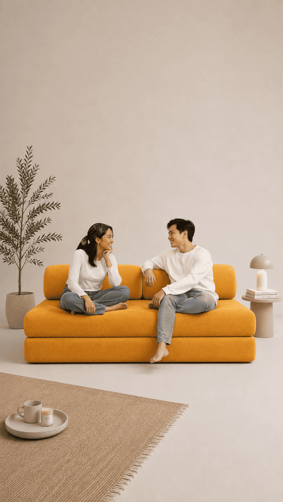 A man and a woman sit on a mustard yellow Sonno Sofa bed in a minimalist beige living room setting, with a potted plant and a small side table in the background.