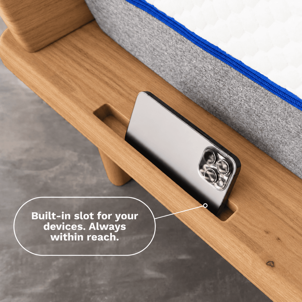 Close-up of a Sonno Signature wooden bed frame with a built-in slot holding a smartphone, with a caption reading 'Built-in slot for your devices. Always within reach.