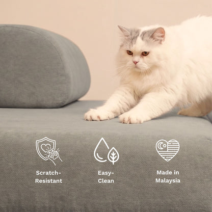 A fluffy white and gray cat lies on a gray upholstered sofa cushion with a matching round pillow behind it. White text icons at the bottom highlight product features: scratch-resistant, easy-clean, and made in Malaysia.