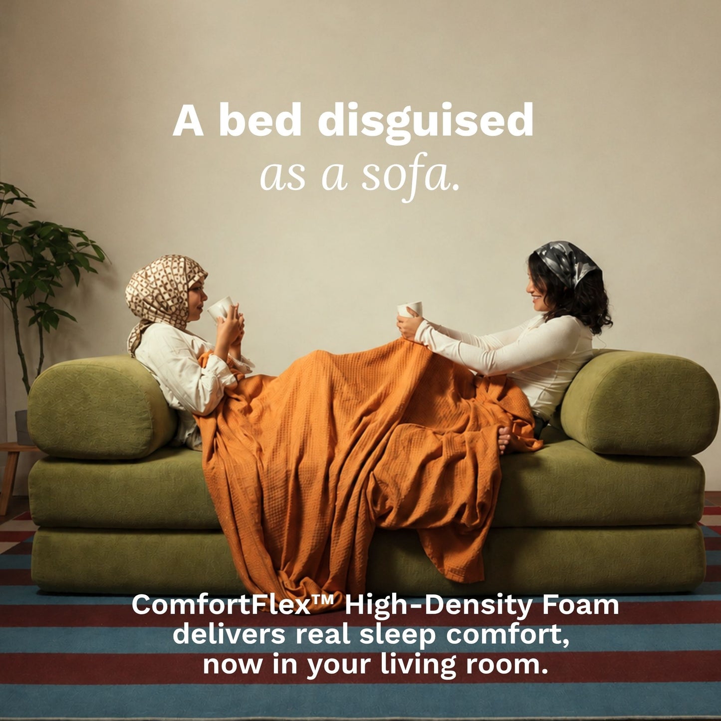 Two women reclining on a green upholstered sofa, covered with an orange blanket, in a living room setting. White text at the top reads 'A bed disguised as a sofa.