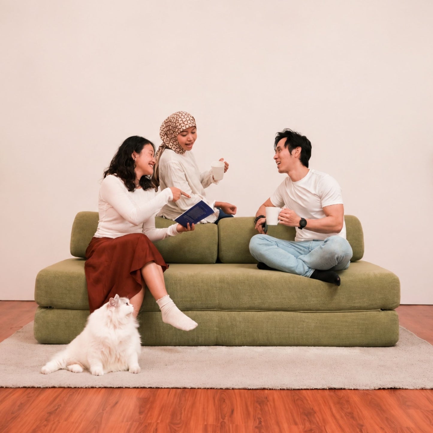 Three people sitting and interacting on a low olive green sofa in a minimalist room, with a white cat on a rug in the foreground. The product appears to be the sofa, shown in a real interior setting with people and an animal present, so this is Lifestyle rather than Product/Packaging.