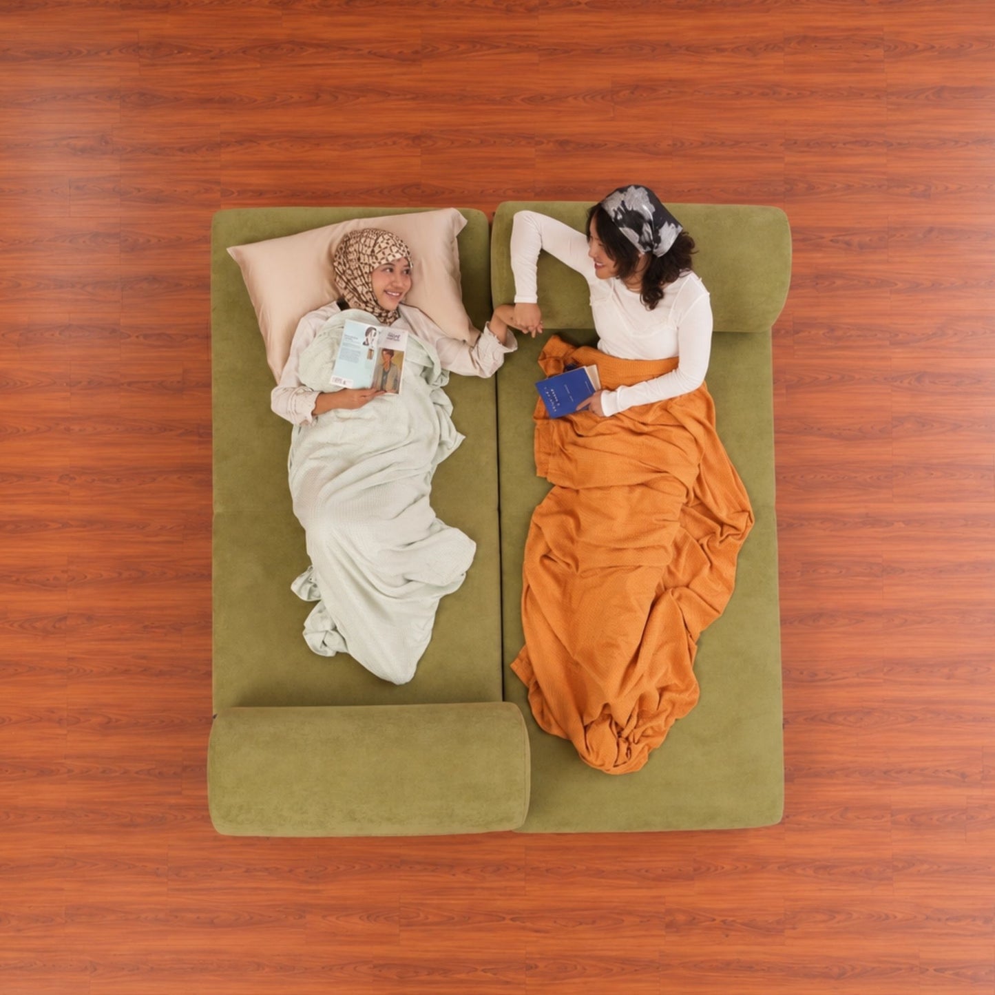 An overhead view of two women sitting on a green sectional sofa, holding hands and reading books under blankets, set against a wood-look floor.