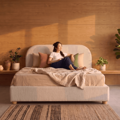 Woman sitting on a Sunset Bed Frame in a cosy bedroom with wooden walls and plants.