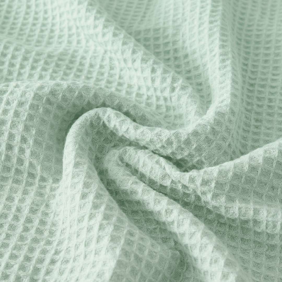 Close-up of a Sonno Waflfe blanket in a green color. The fabric is gathered into soft, heavy folds in the center, highlighting the depth of the "waffle" pockets. The lighting creates subtle shadows within each tiny square