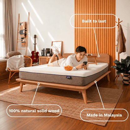A person lies on a mattress on a wooden Sonno Signature bed frame in a styled bedroom. White callout lines with text highlight features: 'Built to last,' '100% natural solid wood,' and 'Made in Malaysia.