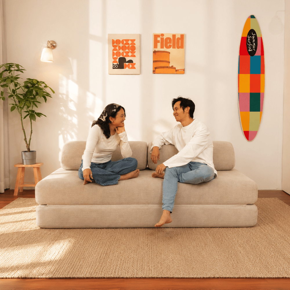 A man and a woman sit on a beige Sonno sofa bed in a bright, modern living room. The room features a potted plant, a small wooden stool, two framed art prints on the wall, and a colorful surfboard decoration.