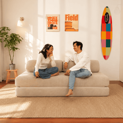 A man and a woman sit on a beige Sonno sofa bed in a bright, modern living room. The room features a potted plant, a small wooden stool, two framed art prints on the wall, and a colorful surfboard decoration.