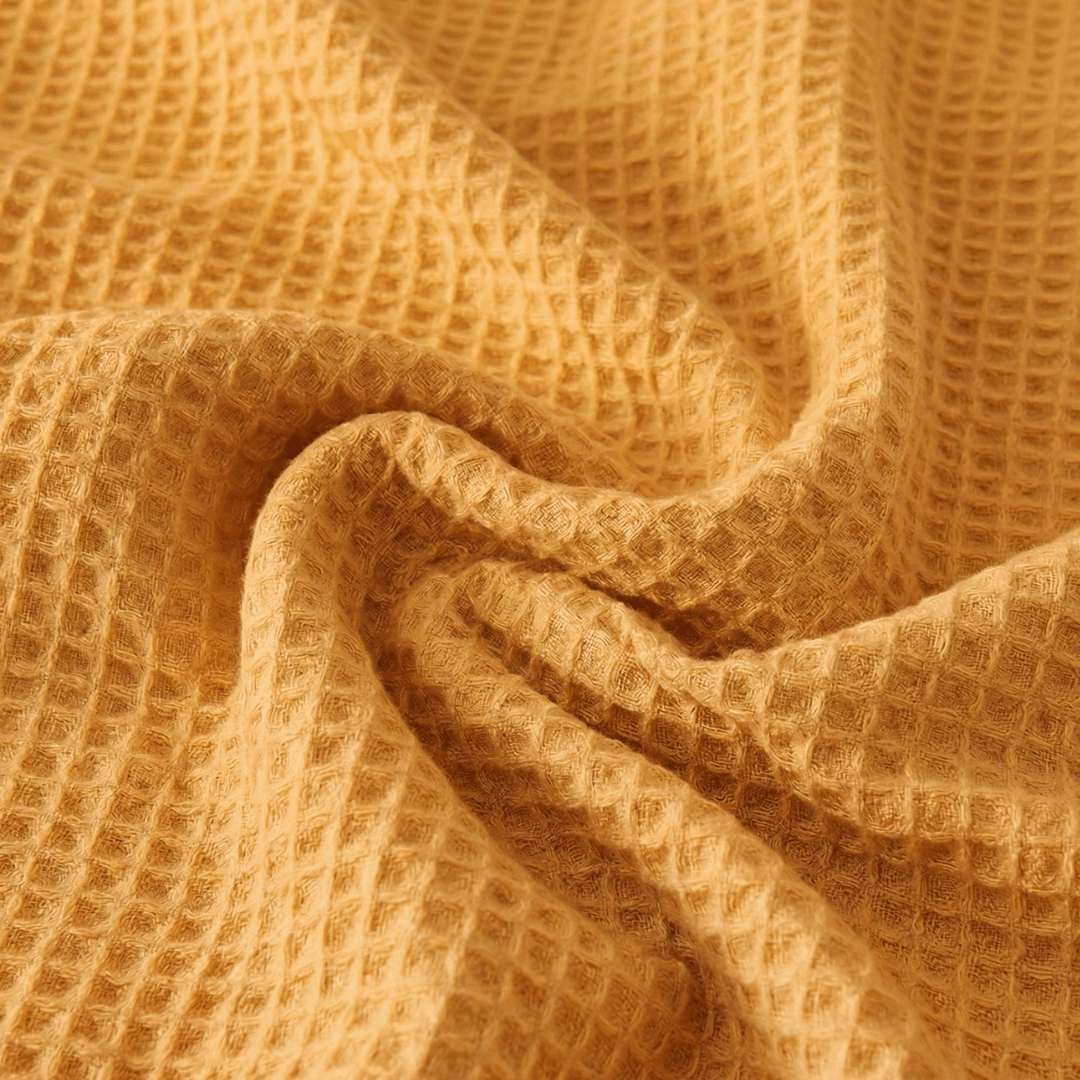 Close-up of a Sonno Waflfe blanket in a warm, mustard-orange or golden-yellow hue. The fabric is gathered into soft, heavy folds in the center, highlighting the depth of the "waffle" pockets. The lighting creates subtle shadows within each tiny square