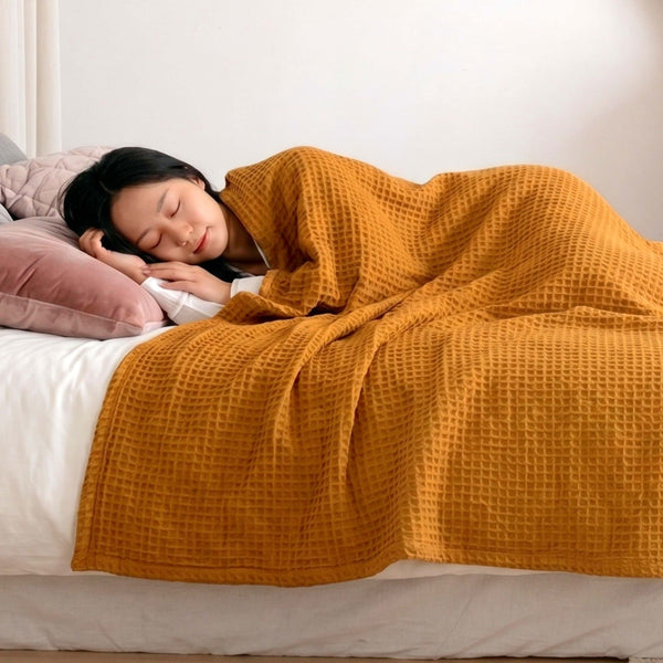 Person sleeping under a mustard yellow waffle blanket on a bed with pink pillows.