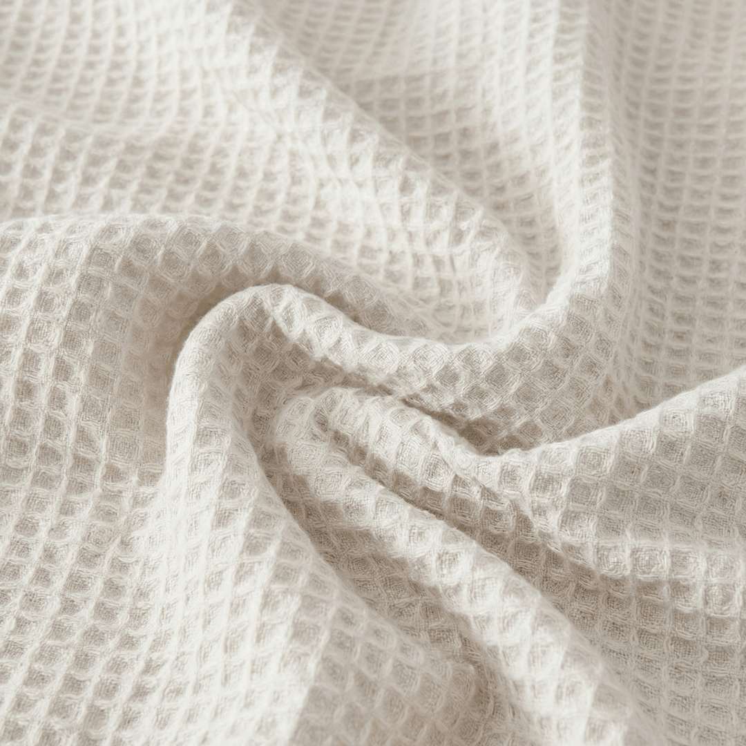 Close-up of a Sonno Waflfe blanket in a white color. The fabric is gathered into soft, heavy folds in the center, highlighting the depth of the "waffle" pockets. The lighting creates subtle shadows within each tiny square