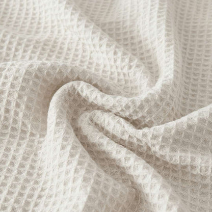 Close-up of a Sonno Waflfe blanket in a white color. The fabric is gathered into soft, heavy folds in the center, highlighting the depth of the "waffle" pockets. The lighting creates subtle shadows within each tiny square