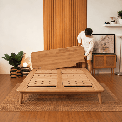 A man in a white tracksuit is assembling a light oak Sonno Signature wooden platform bed frame in a modern, warm-toned room with wood paneling and a woven rug.