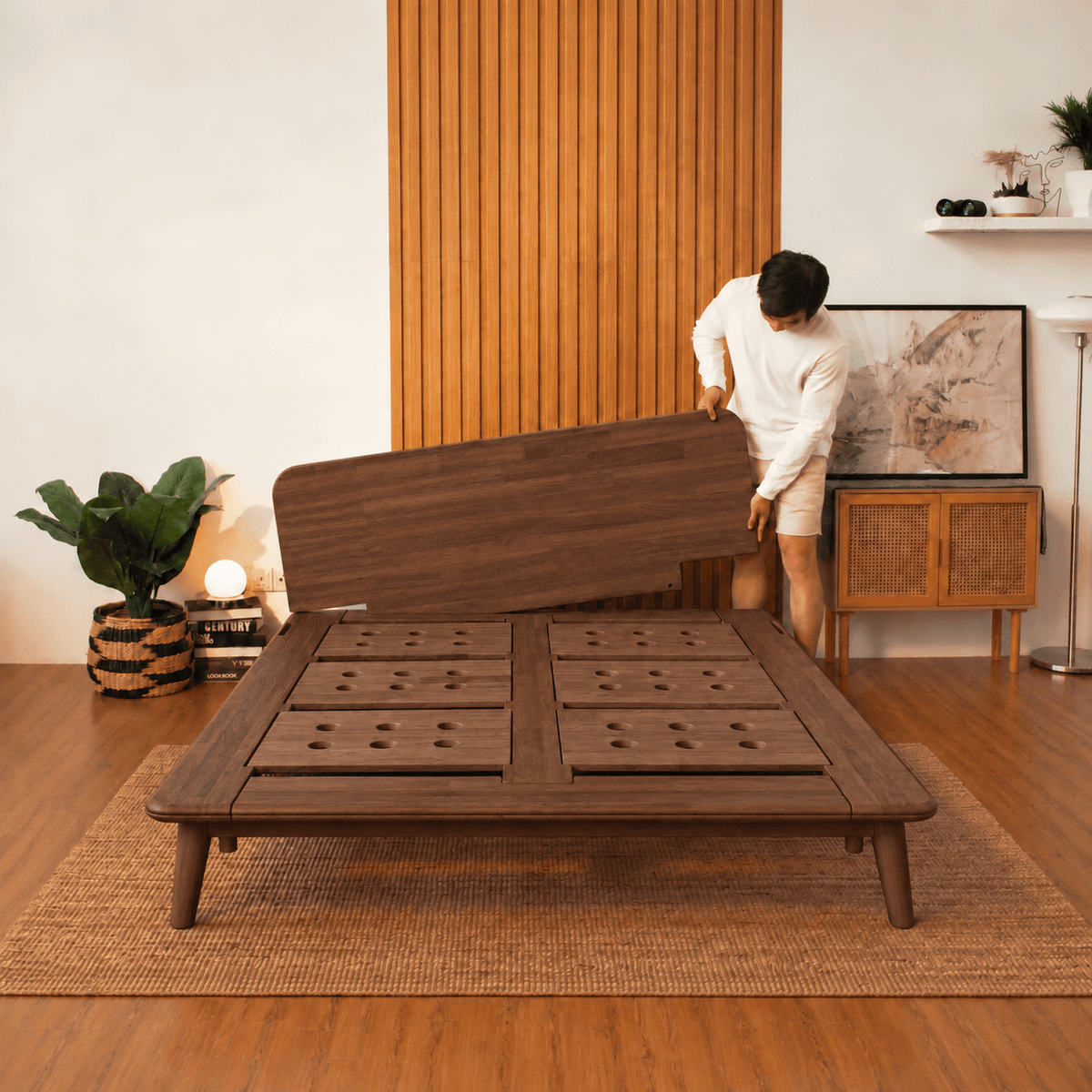 A man in a white outfit is assembling a dark brown walnut Sonno Signature wooden low-profile platform bed frame in a modern, warm-toned room with wood paneling and a woven rug.
