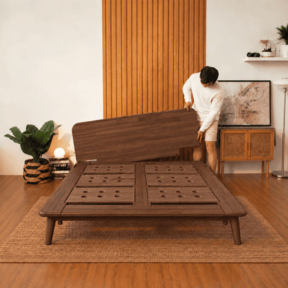 A man in a white outfit is assembling a dark brown walnut Sonno Signature wooden low-profile platform bed frame in a modern, warm-toned room with wood paneling and a woven rug.