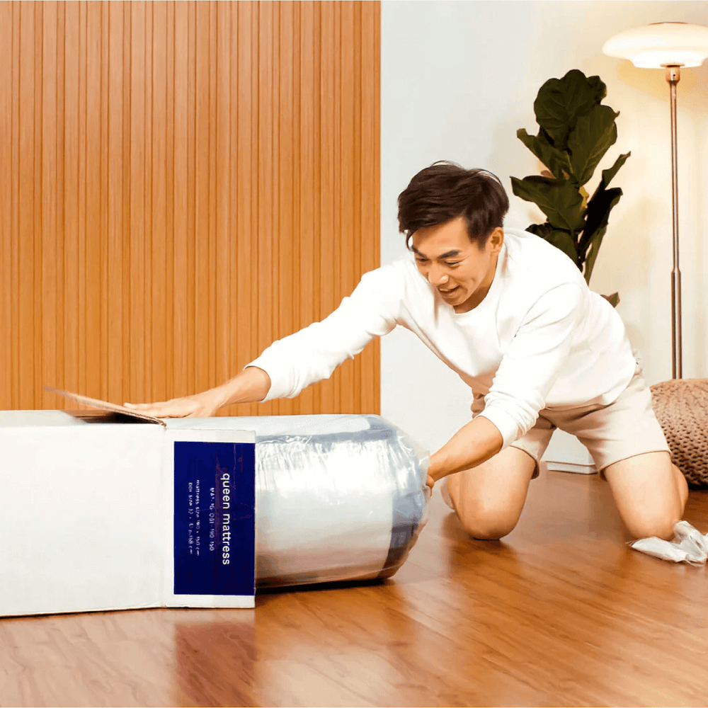 Image of a person excitedly unboxing the Sonno Original Mattress. The individual is kneeling beside the packaging, removing protective layers from the rolled mattress. The setting features a cozy room with wooden flooring and plants, highlighting the ease of setup and the anticipation of a comfortable sleep experience.